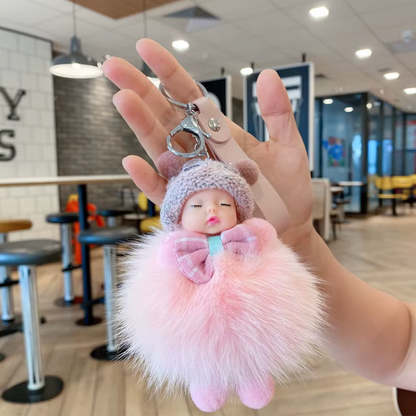 Keychain with a small doll figure and pink fluffy ball held in a hand indoors.