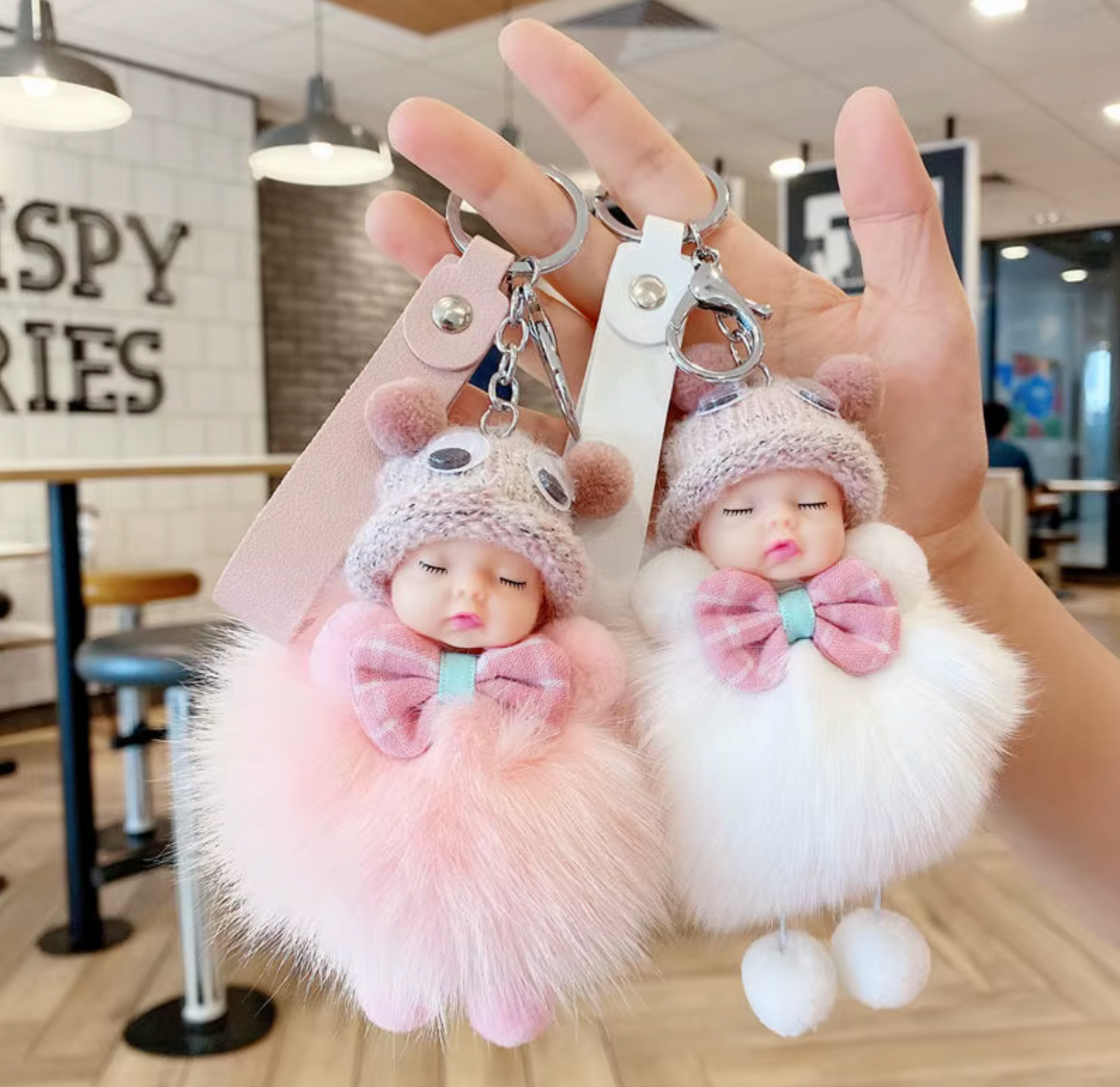 Two fluffy keychains shaped like dolls with bows, held in a hand indoors.