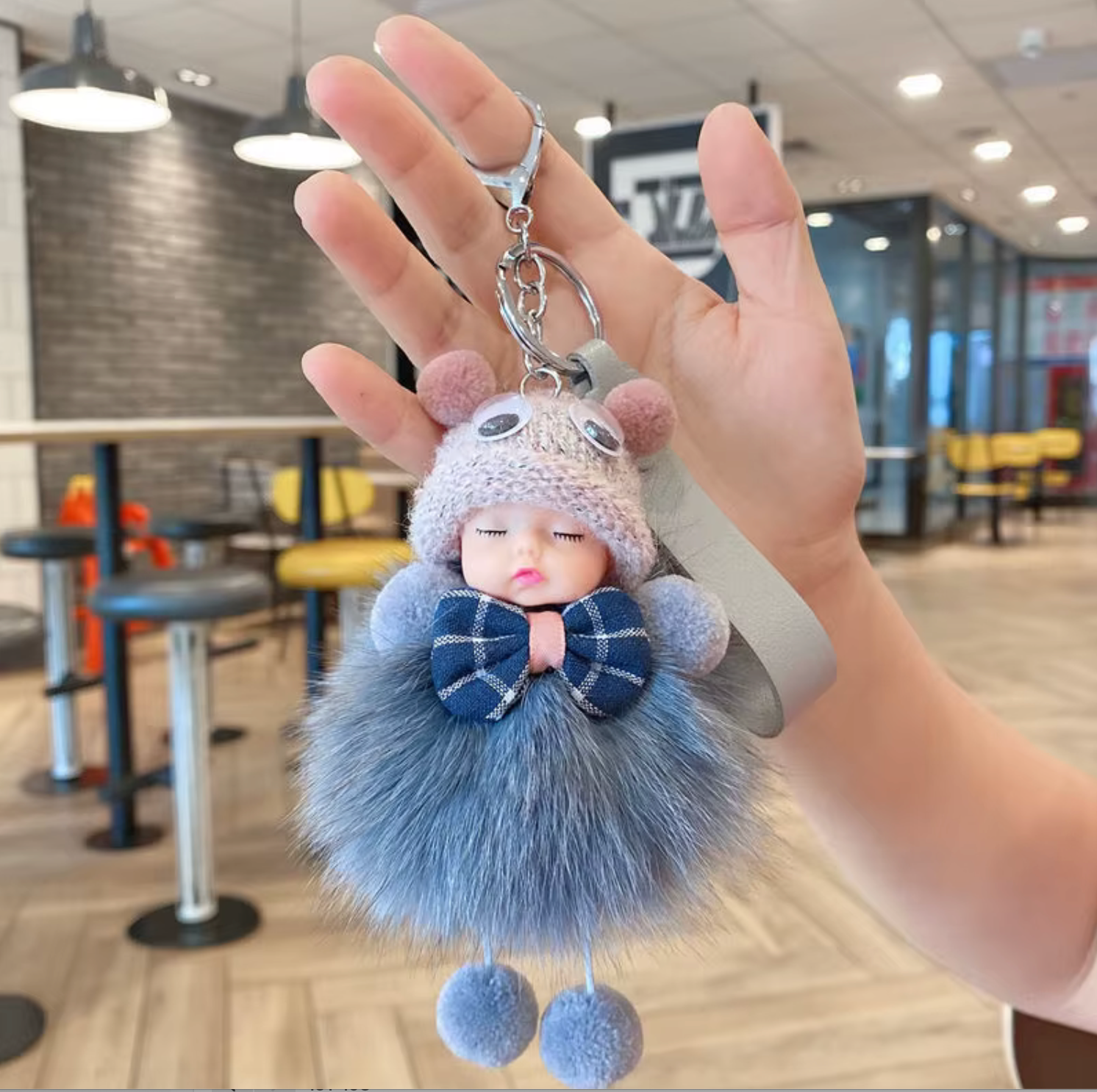 Fuzzy keychain with a doll-like figure held in a hand indoors.