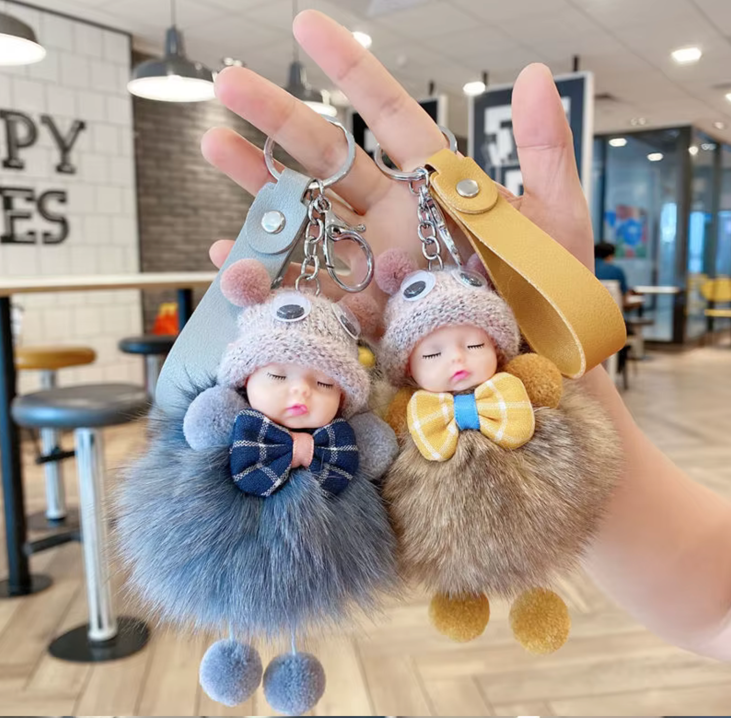 Two fluffy keychains shaped like sleeping babies with bow ties, held in a hand indoors.