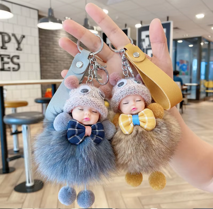 Two fluffy keychains shaped like sleeping babies with bow ties, held in a hand indoors.