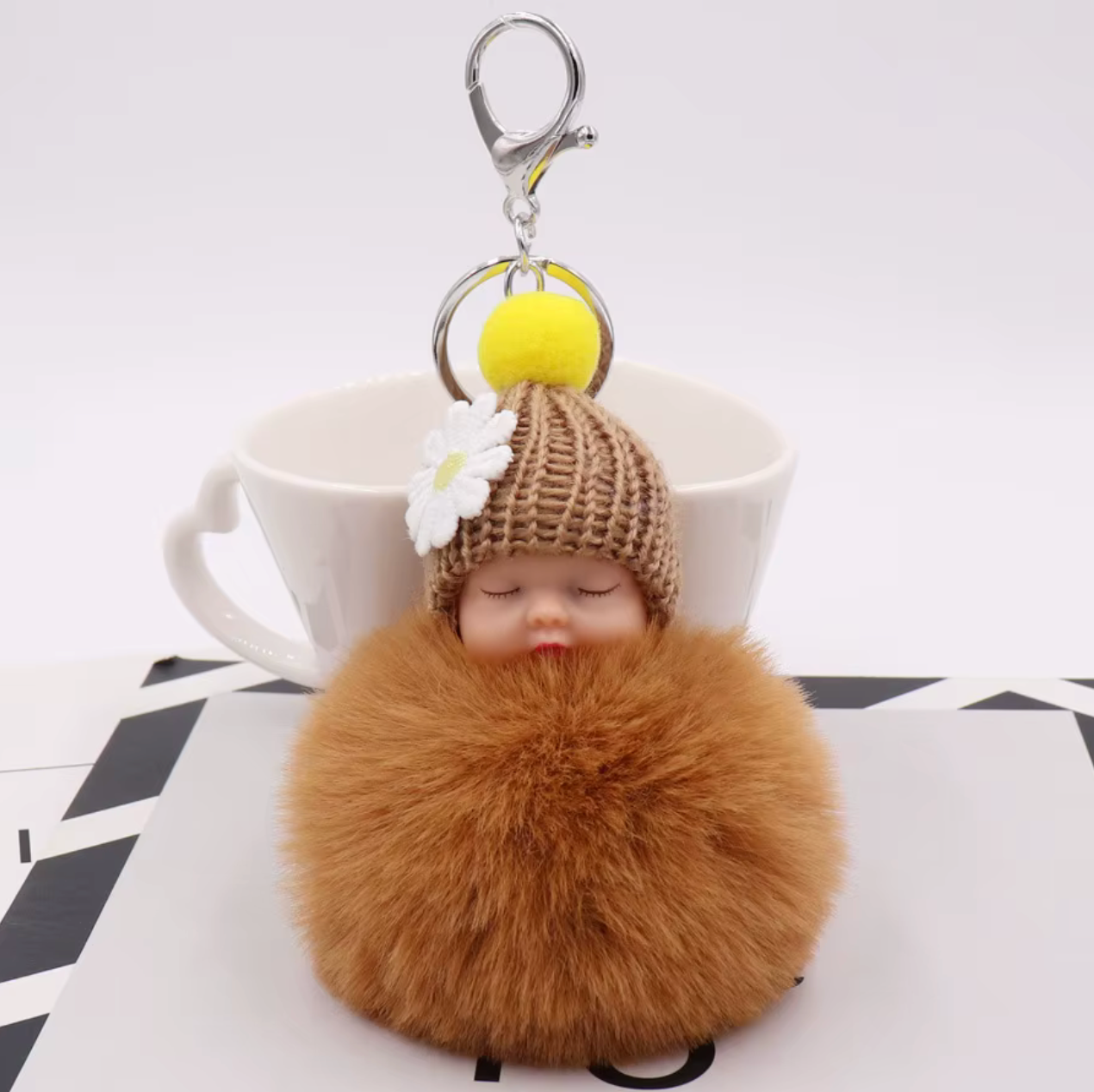 Brown fluffy keychain with a small doll wearing a knitted hat and flower, against a white background.