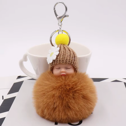 Brown fluffy keychain with a small doll wearing a knitted hat and flower, against a white background.