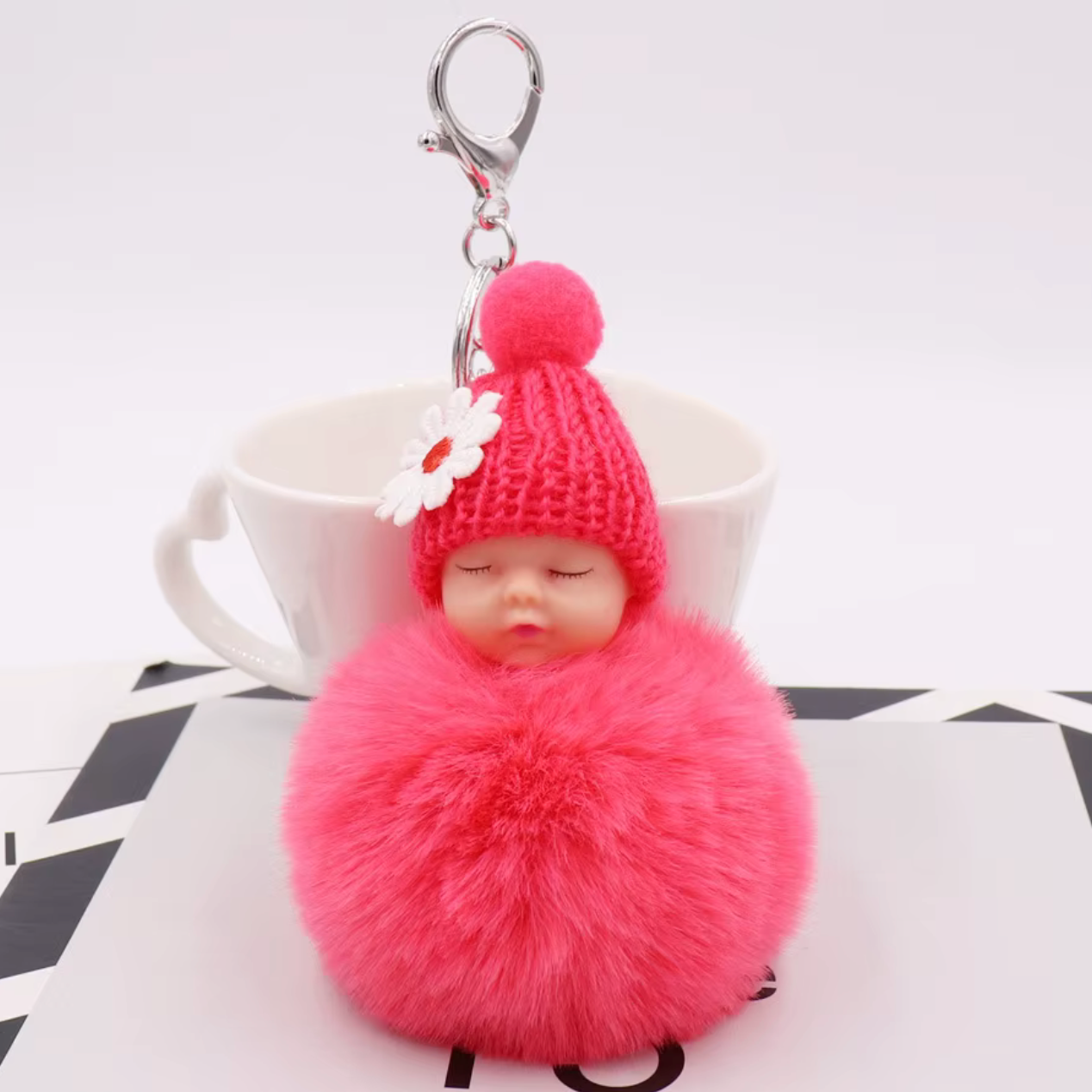 Pink fluffy keychain with a small doll wearing a knitted hat on a white surface.