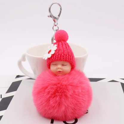 Pink fluffy keychain with a small doll wearing a knitted hat on a white surface.