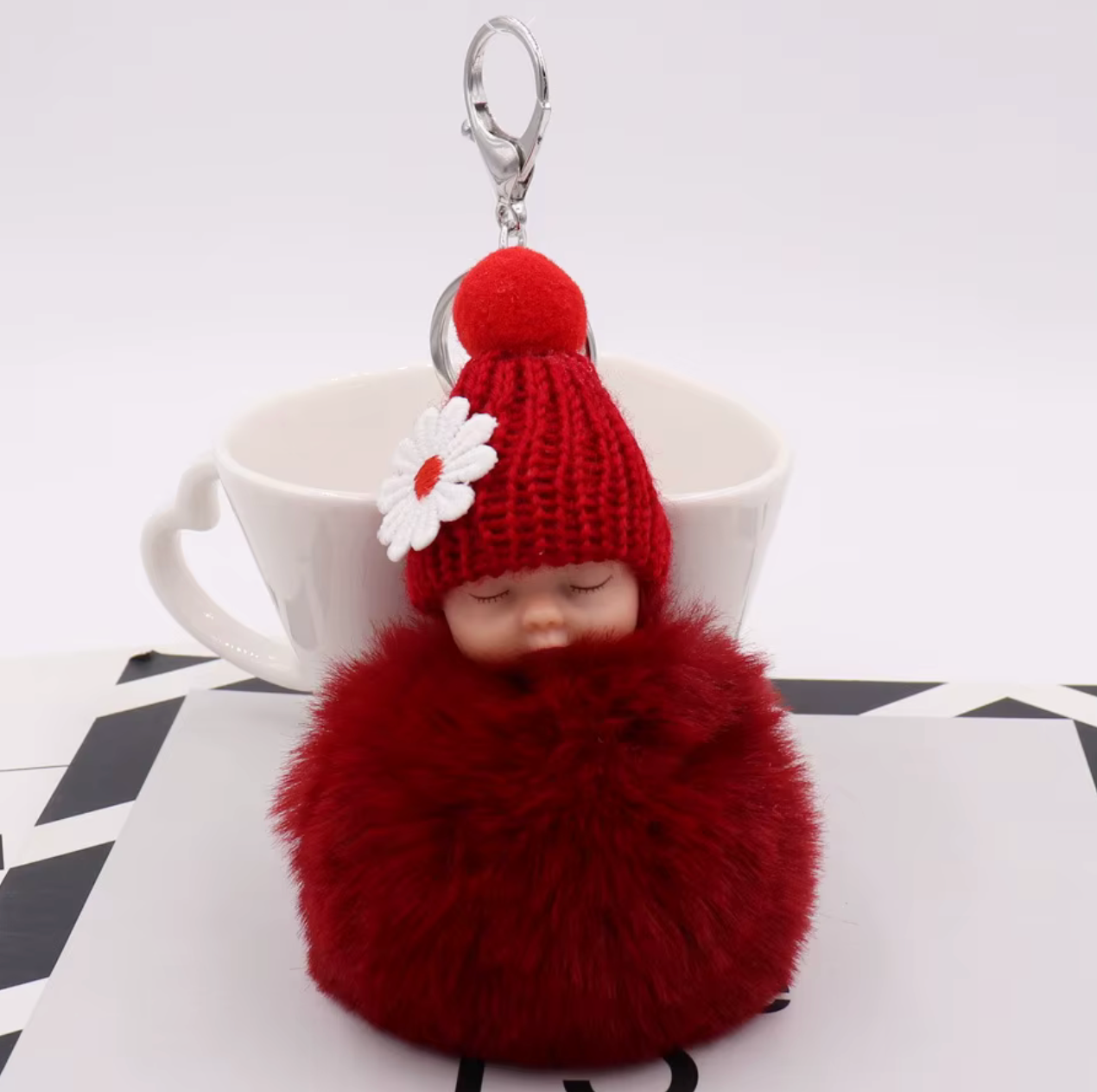 Red fluffy keychain with a small doll wearing a knitted hat on a white surface.