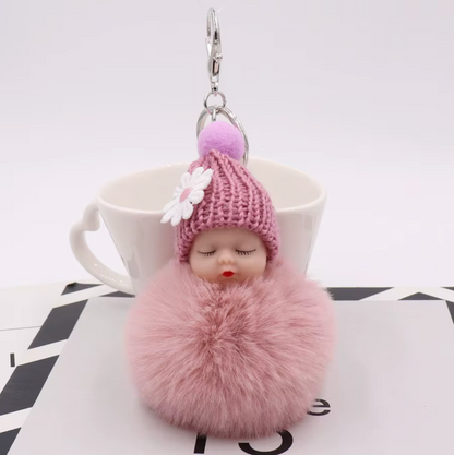 Pink fluffy keychain with a small doll-like figure on top, placed on a white surface.
